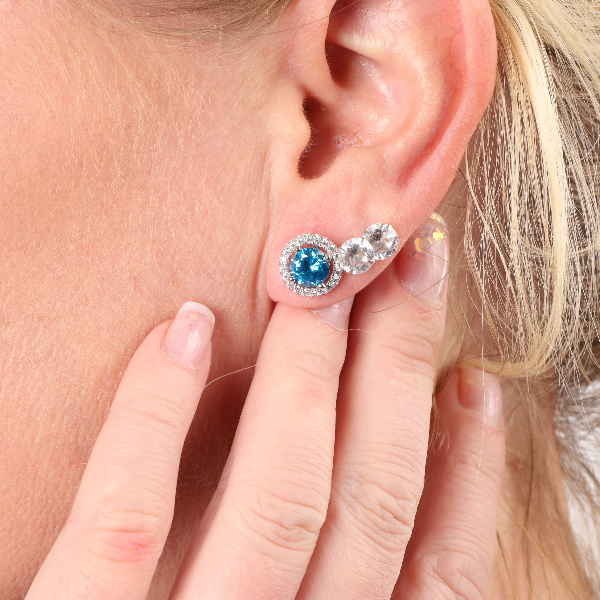 Close-up of a person's ear adorned with the Blue Horizon Radiant Studs, featuring sparkling blue and clear gemstones, delicately touched by fingers—Mia Ishaaq's sterling silver earrings enhancing the elegance.