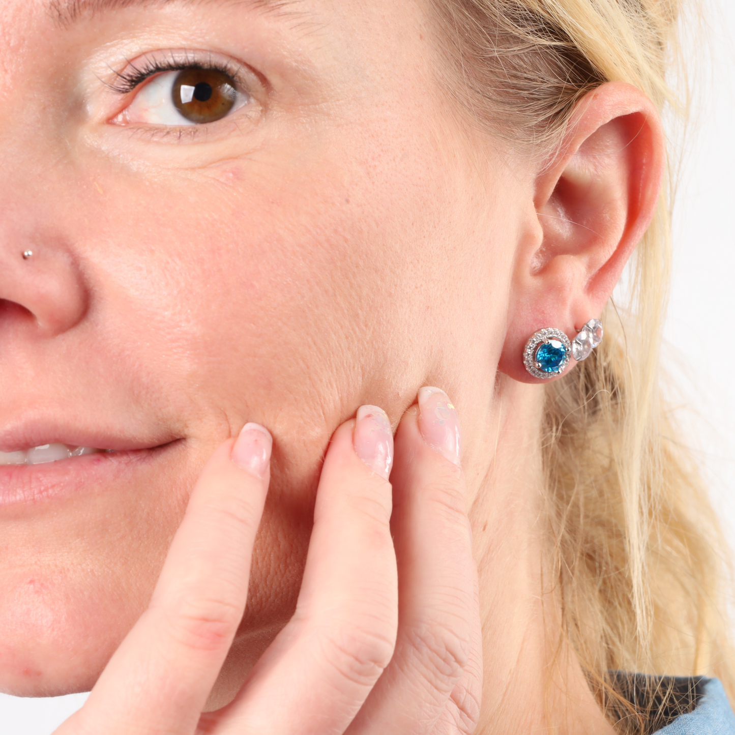 A woman wearing the Blue Horizon Radiant Studs by Mia Ishaaq gently touches her cheek, showing skincare concern or examination.