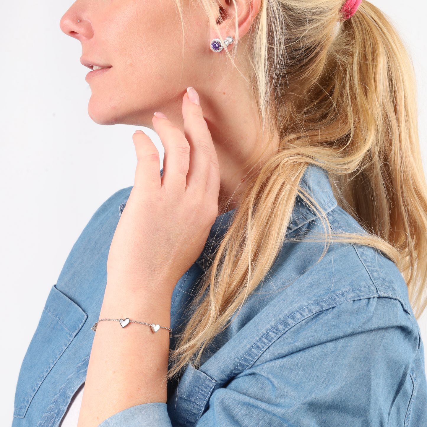 A woman in a denim shirt and ponytail wears a heart bracelet and gem earrings, her fingers gently touching her neck. Her elegance is highlighted by the shimmer of the VIOLET CZ Stud Earrings by Mia Ishaaq, crafted from 925 sterling silver.