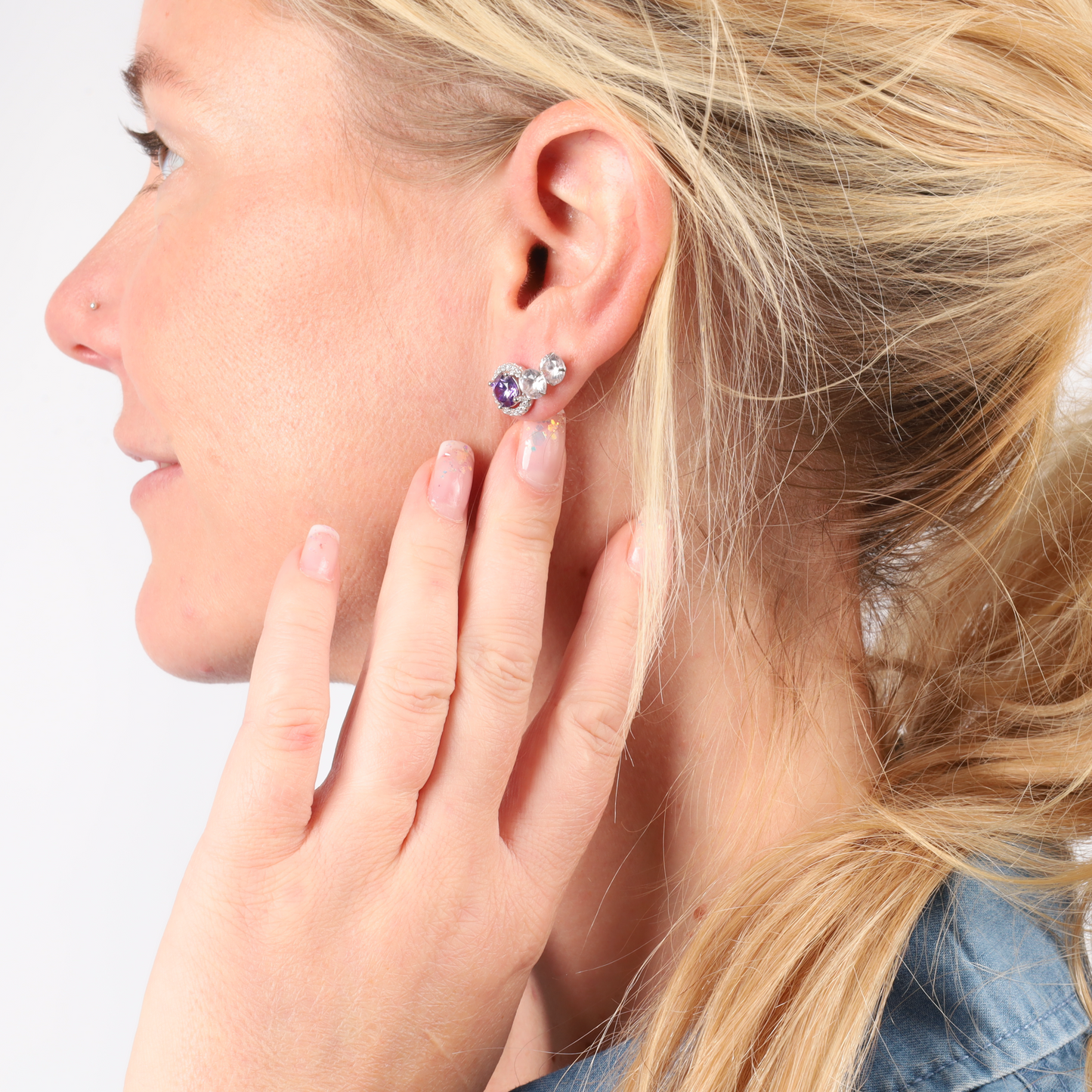 A woman with light blonde hair draped over a denim jacket is shown in profile, touching her ear adorned with Mia Ishaaq's 925 Sterling Silver VIOLET CZ Stud Earrings.