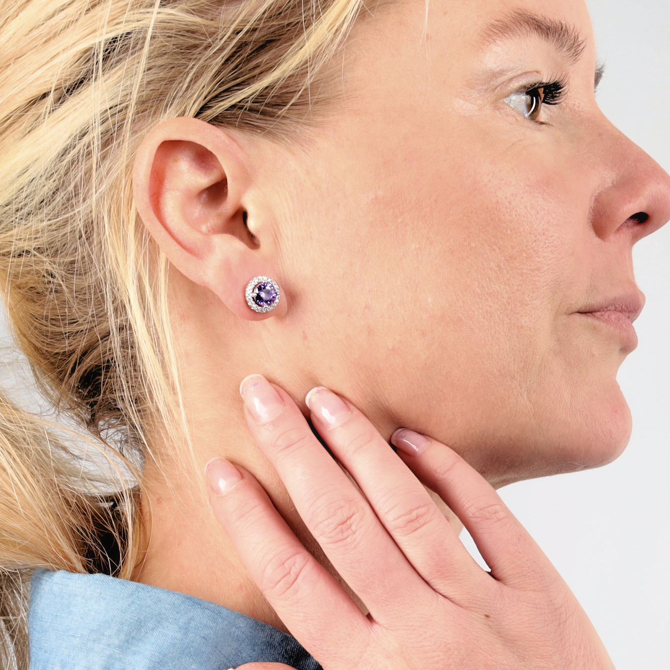 A woman with blond hair, adorned in the Mia Ishaaq 925 Sterling Silver VIOLET CZ Stud Earrings, touches her neck and gazes to the right against a light background.