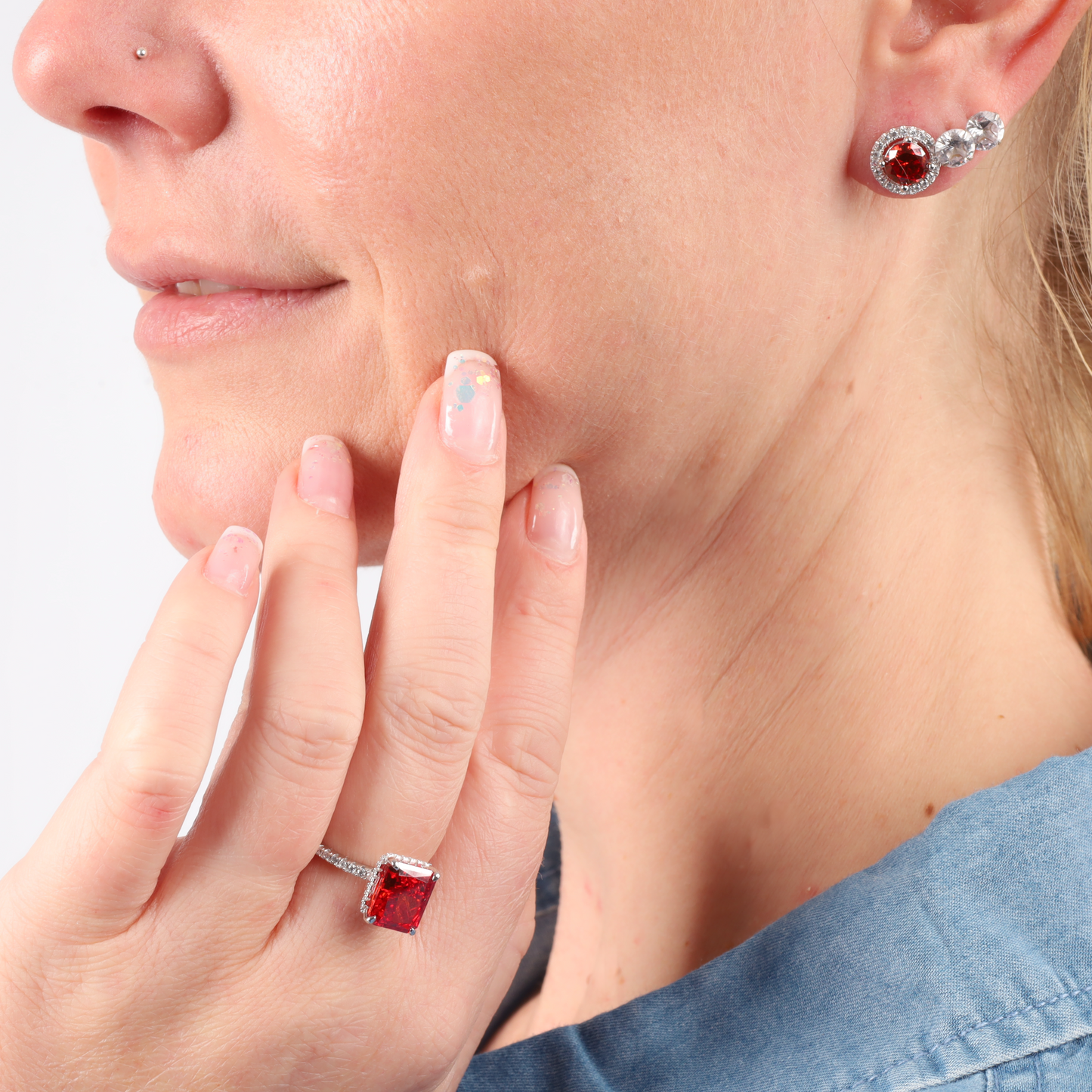 A woman touches her cheek, adorned with the Mia Ishaaq Sterling Silver Red Radiant Ring and matching earrings set with red simulant stones.