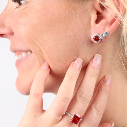 A woman smiles while wearing the Mia Ishaaq Sterling Silver Red Radiant Ring, paired with elegant red simulant stone earrings. Her hand, showcasing the ring's vibrant cubic zirconia stones, is gracefully positioned near her face against a white background.