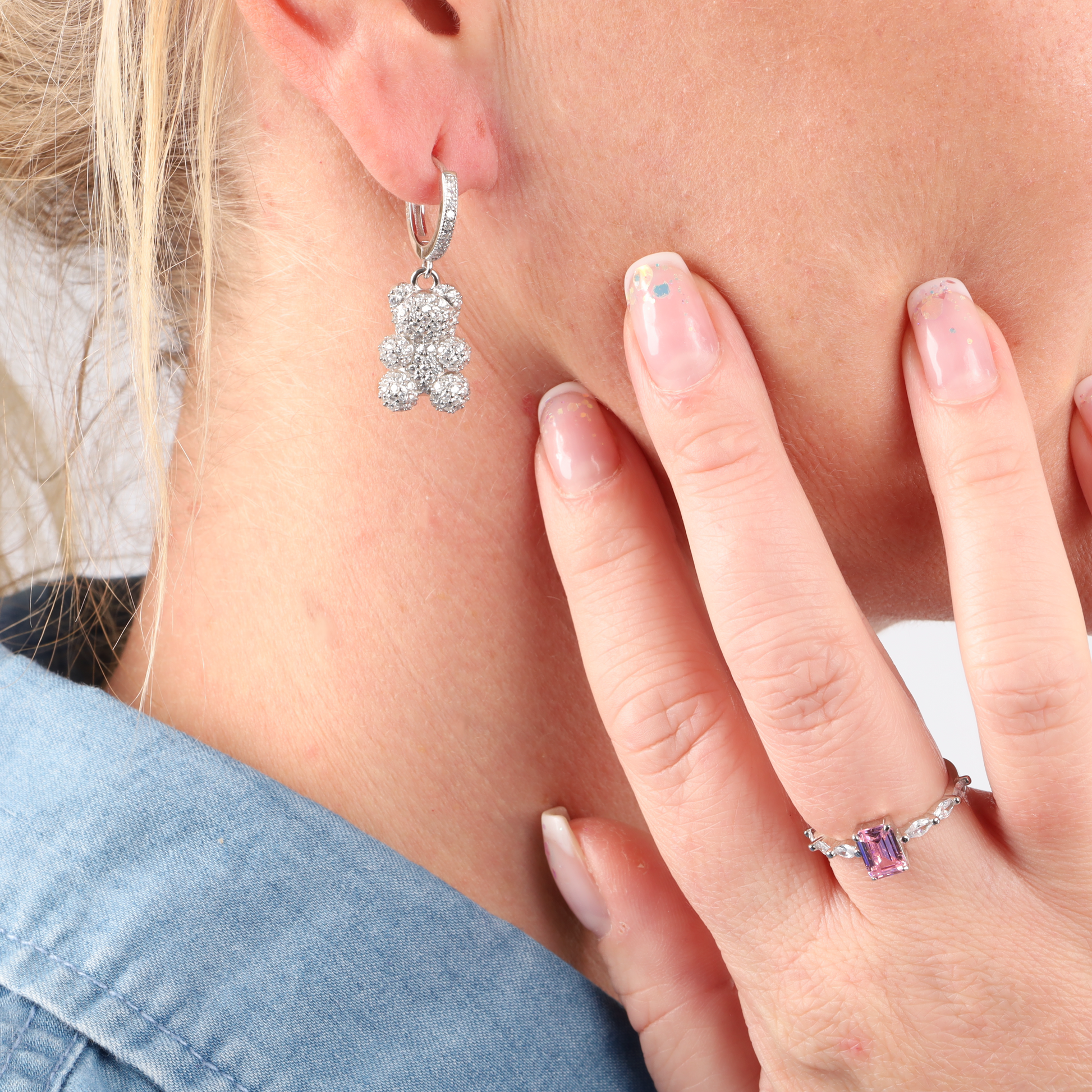 Close-up of a person wearing Mia Ishaaq's Pave Nostalgia Teddy Bear Earrings and a sparkly ring, with their hand gently touching their face, in a denim shirt.