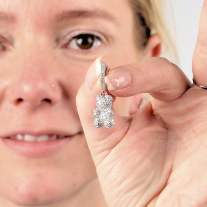 An individual holds a dazzling Pave Nostalgia Teddy Bear Earring by Mia Ishaaq, crafted from sterling silver, close to their eye, highlighting the intricate details of the jewelry.