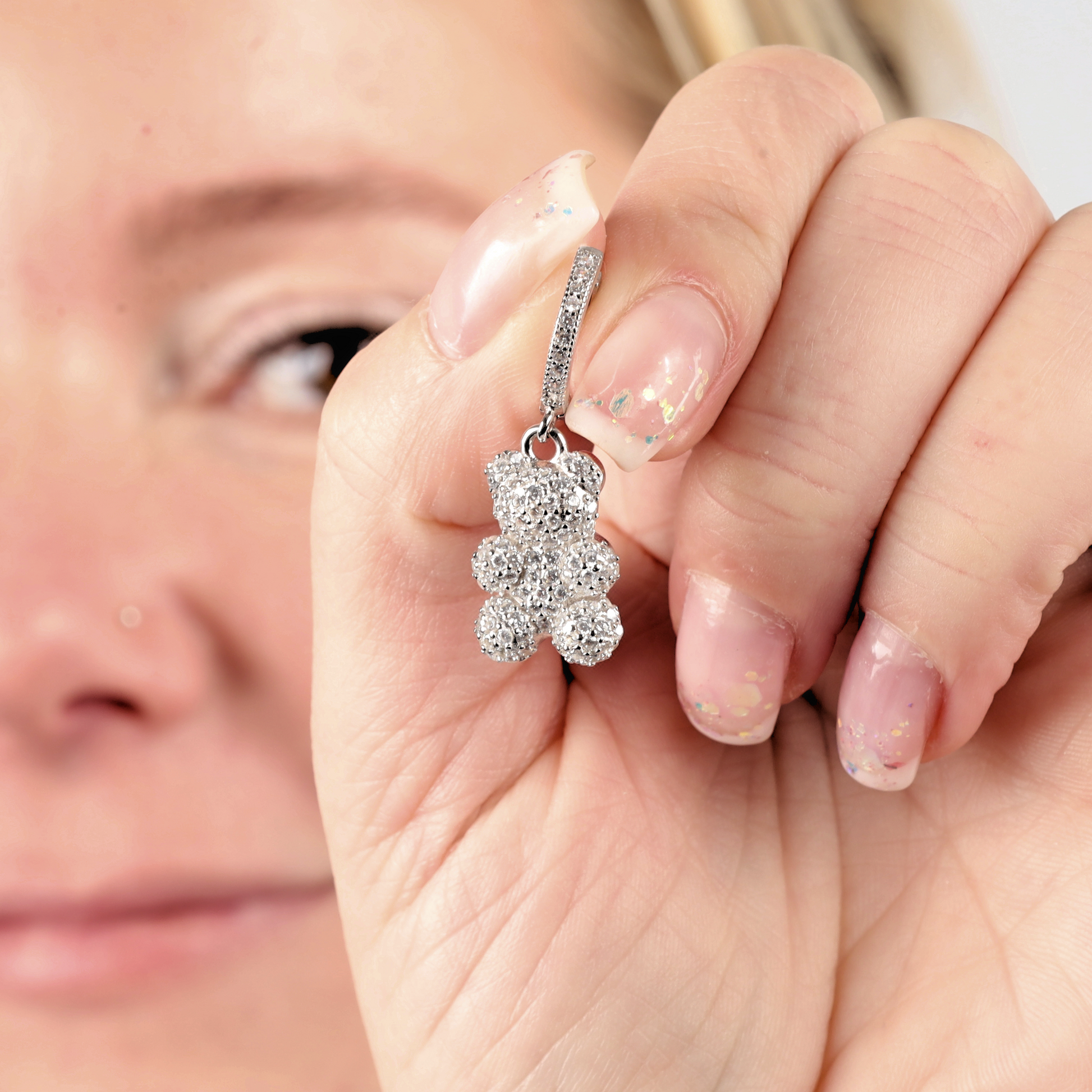 Person holding a shimmering pair of Pave Nostalgia Teddy Bear Earrings by Mia Ishaaq close to their face, their sparkle reminiscent of sterling silver elegance.