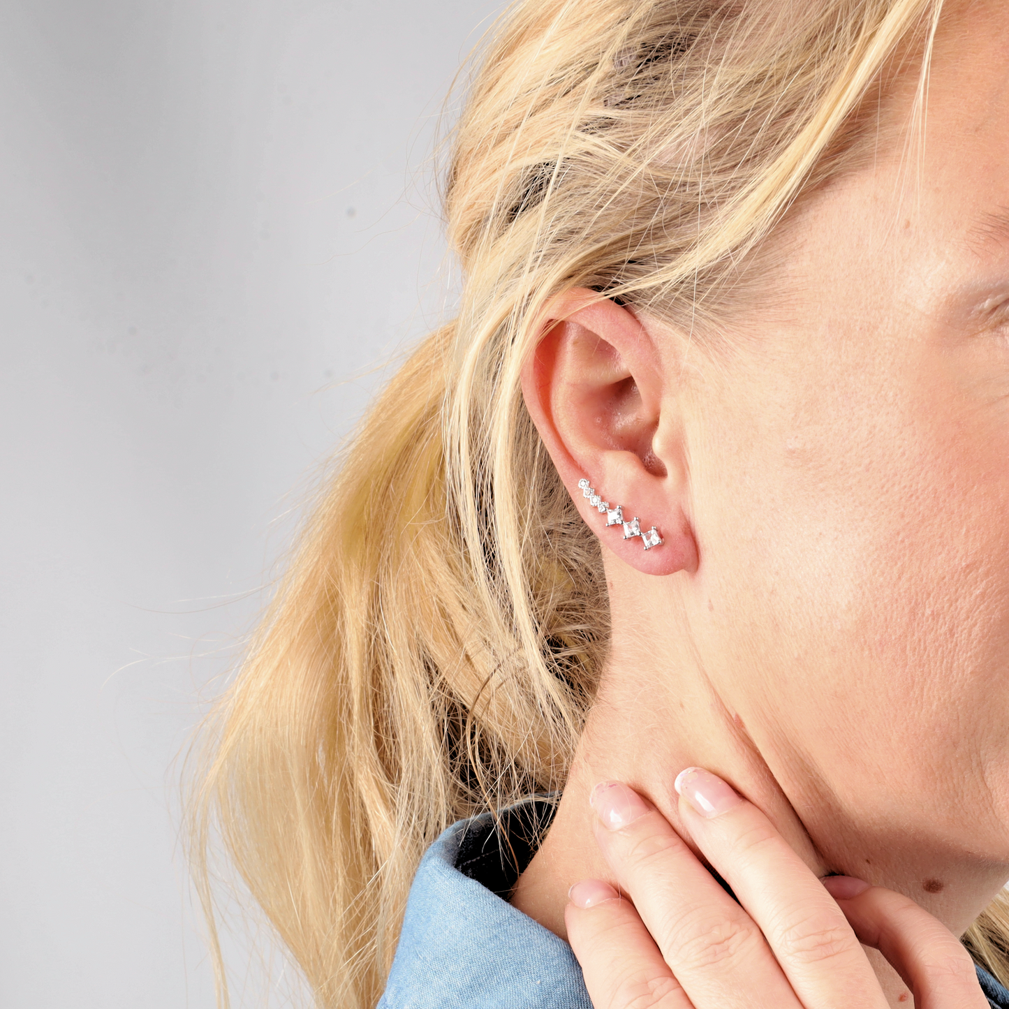 A woman with blonde hair in a ponytail wears Mia Ishaaq's Crystal Ear Cuffs Earrings, gently touching her neck and donning a denim shirt.