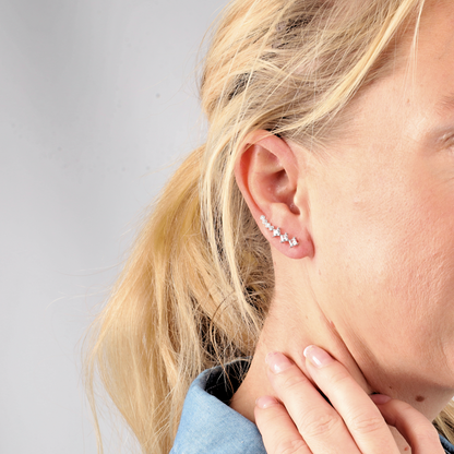 A woman with blonde hair in a ponytail wears Mia Ishaaq's Crystal Ear Cuffs Earrings, gently touching her neck and donning a denim shirt.