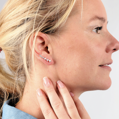 A woman with blonde hair is seen wearing star-shaped earrings along with the Mia Ishaaq Crystal Ear Cuffs Earrings, styled with a blue shirt. She touches her ear and smiles radiantly.