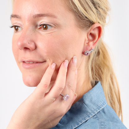 A woman in a denim shirt gently touches her face, adorned with Mia Ishaaq's 925 Sterling Silver PINK CZ Stud Earrings and a matching ring, each piece set in elegant sterling silver.