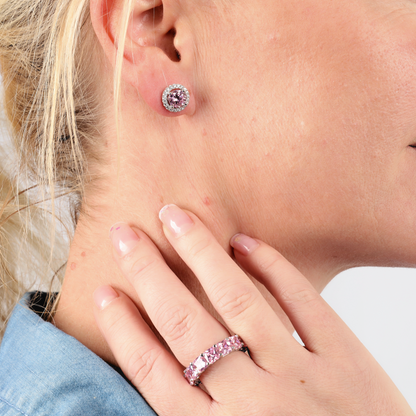A close-up captures a person adorned with bold jewellery, featuring pink simulant stone earrings and a matching Mia Ishaaq Sterling Silver Pink Wide Band Ring, gently touching their neck.