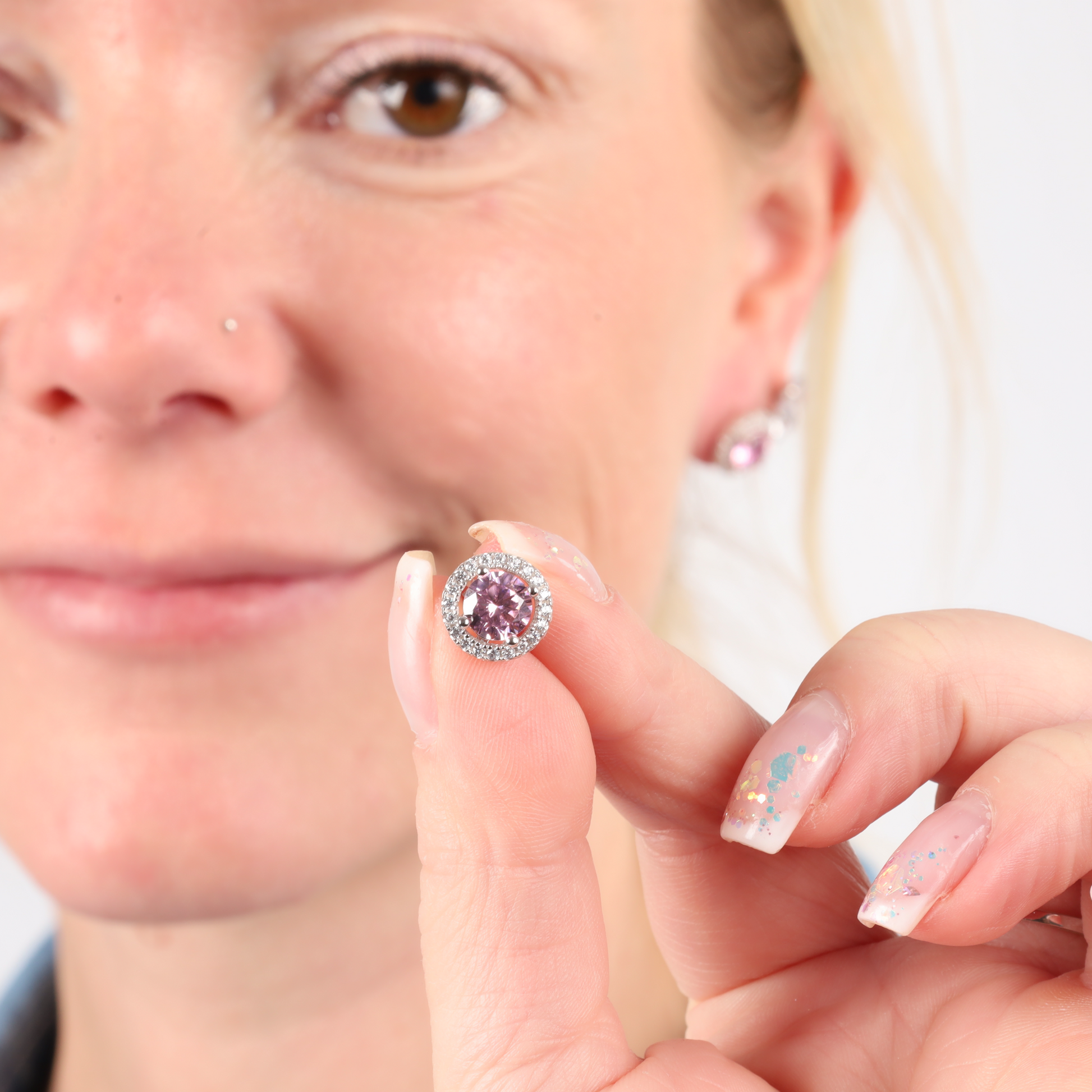 A person holding a round pink gemstone near their face, complemented by Mia Ishaaq's 925 Sterling Silver PINK CZ Stud Earrings and matching nail polish.