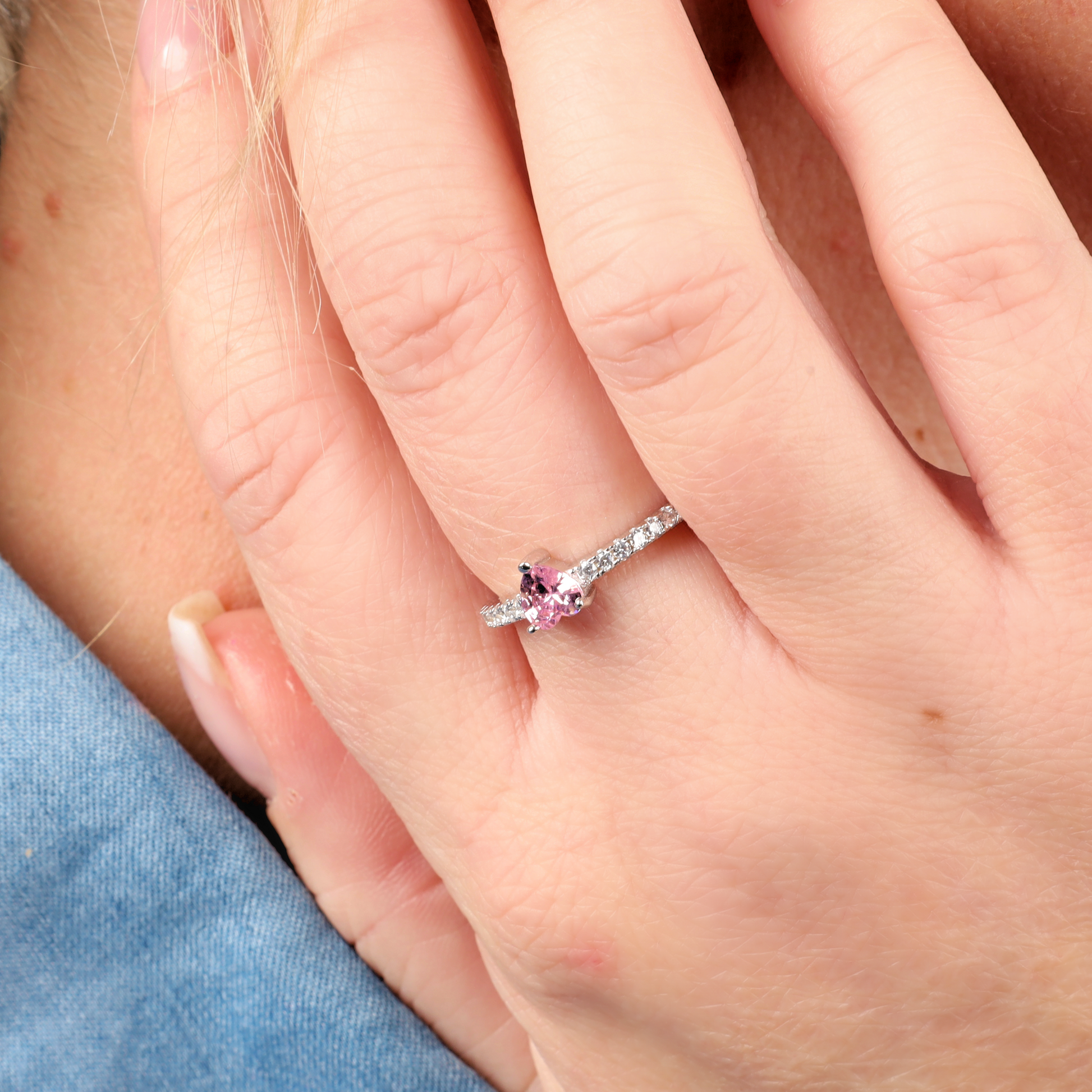 Displayed in the photo is a close-up of a hand wearing the Sterling Silver Heart Solitaire Ring from Mia Ishaaq. The ring showcases a pink cubic zirconia gemstone accented by sparkling diamond details, set against a lush blue fabric backdrop.