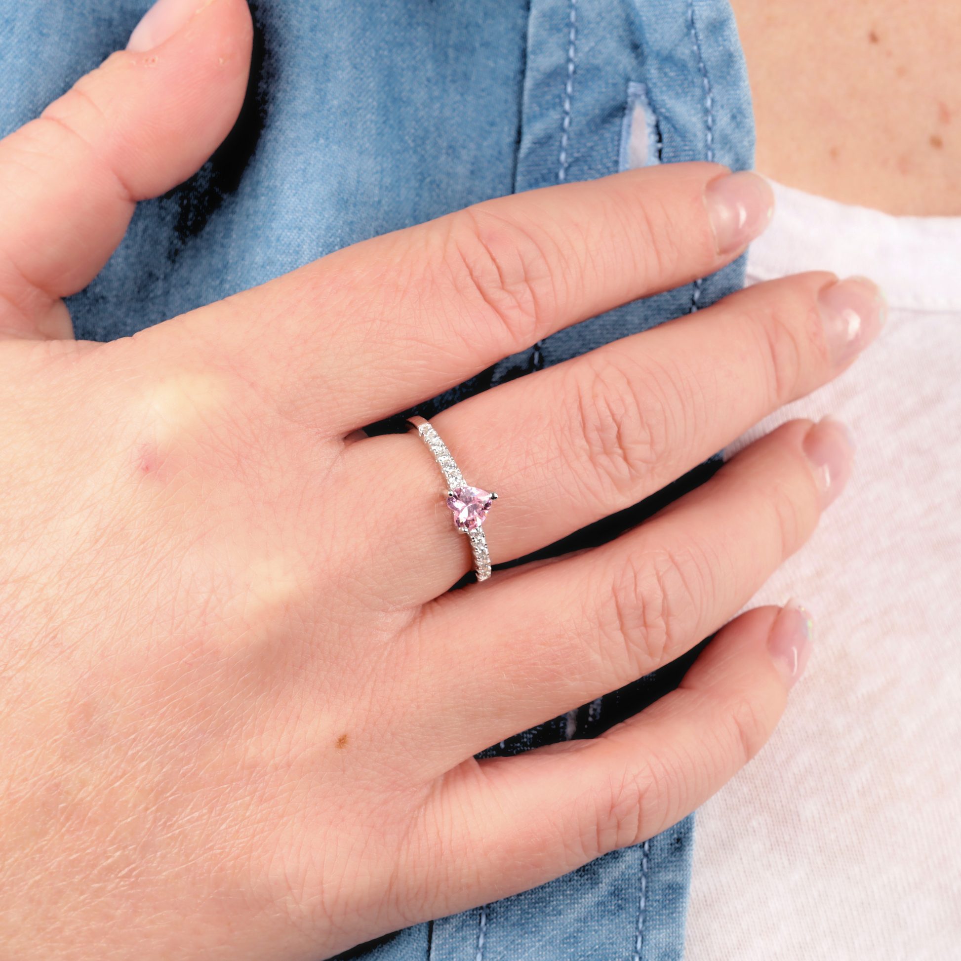 A hand adorned with the Mia Ishaaq Sterling Silver Heart Solitaire Ring, featuring a stunning pink cubic zirconia and delicate diamonds, elegantly rests on a denim-clad shoulder.