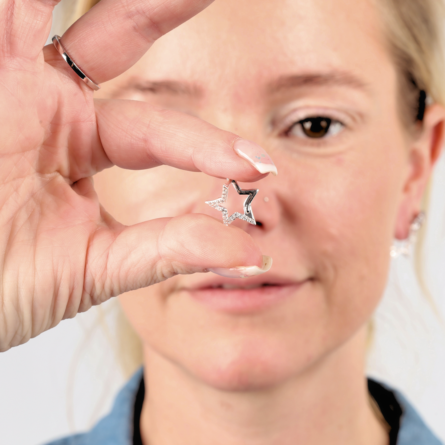 A person delicately holds a small star-shaped earring made of 925 sterling silver between their fingers, its surface twinkling like the Sparkly Star Huggie Hoop Earrings by Mia Ishaaq, perfectly catching the light in front of their face.