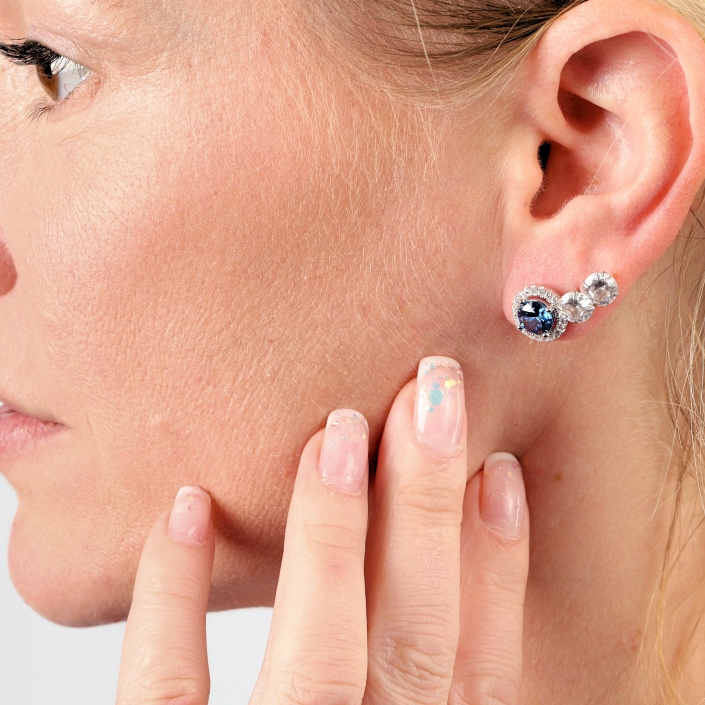 A close-up of a woman touching her ear highlights the Blue Lagoon Radiance Studs by Mia Ishaaq, sterling silver earrings set with sapphire blue CZ stones and clear accents. Enhanced with white gold plating, these elegant earrings beautifully catch the light.