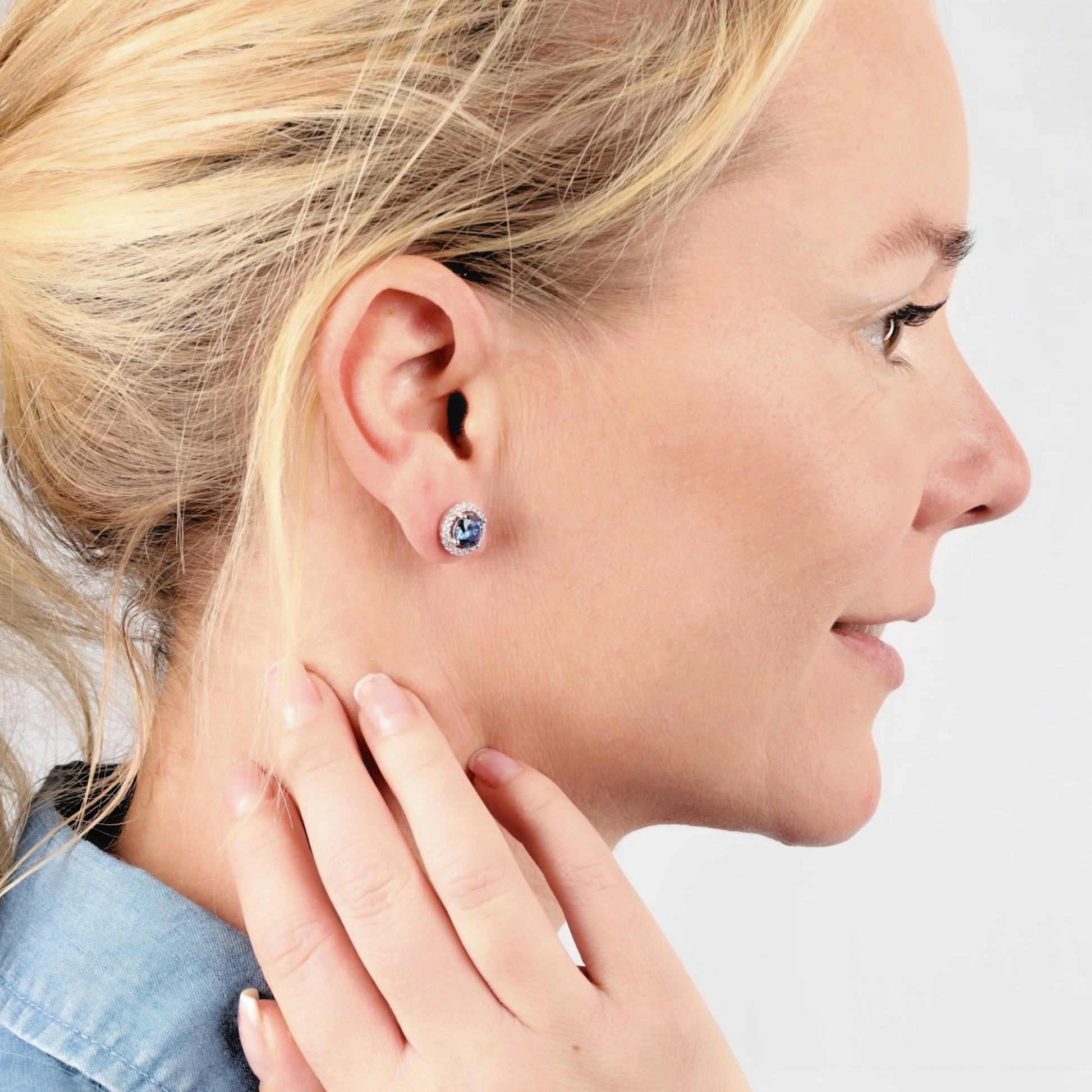 A woman with blonde hair wears the striking Blue Lagoon Radiance Studs by Mia Ishaaq, adorned with white gold plating. She delicately touches her neck, highlighting the elegance of her blue shirt.