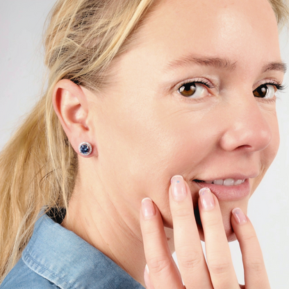 A smiling woman with blonde hair wears the stunning Blue Lagoon Radiance Studs by Mia Ishaaq. She rests her hand on her cheek, showcasing the sparkle of sterling silver and white gold plating against her denim shirt.