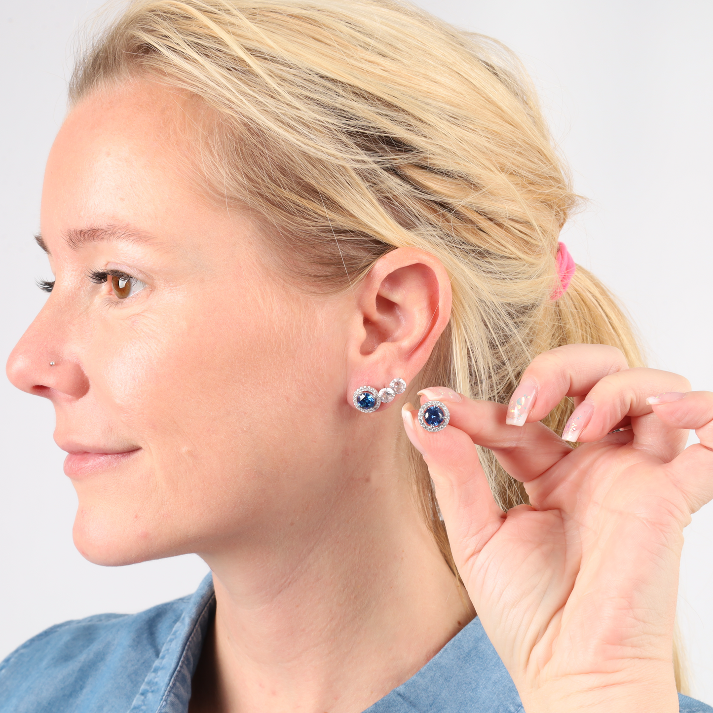 A woman with blonde hair wears the Blue Lagoon Radiance Studs by Mia Ishaaq, their sapphire blue CZ stones set in sterling silver catching the light. In her hand, she holds its match, where a hint of white gold plating glistens; her smile is subtle yet bright.