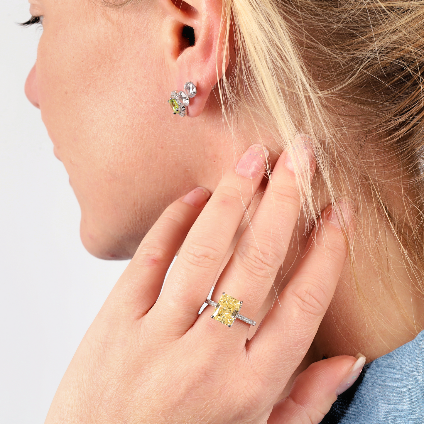 A woman with blonde hair elegantly showcases a Mia Ishaaq Crushed Ice Yellow Radiant Promise Ring on her finger, complemented by matching earrings. The pieces shimmer beautifully, their brilliance enhanced by the white gold plating.