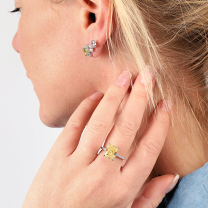 A woman with blonde hair elegantly showcases a Mia Ishaaq Crushed Ice Yellow Radiant Promise Ring on her finger, complemented by matching earrings. The pieces shimmer beautifully, their brilliance enhanced by the white gold plating.