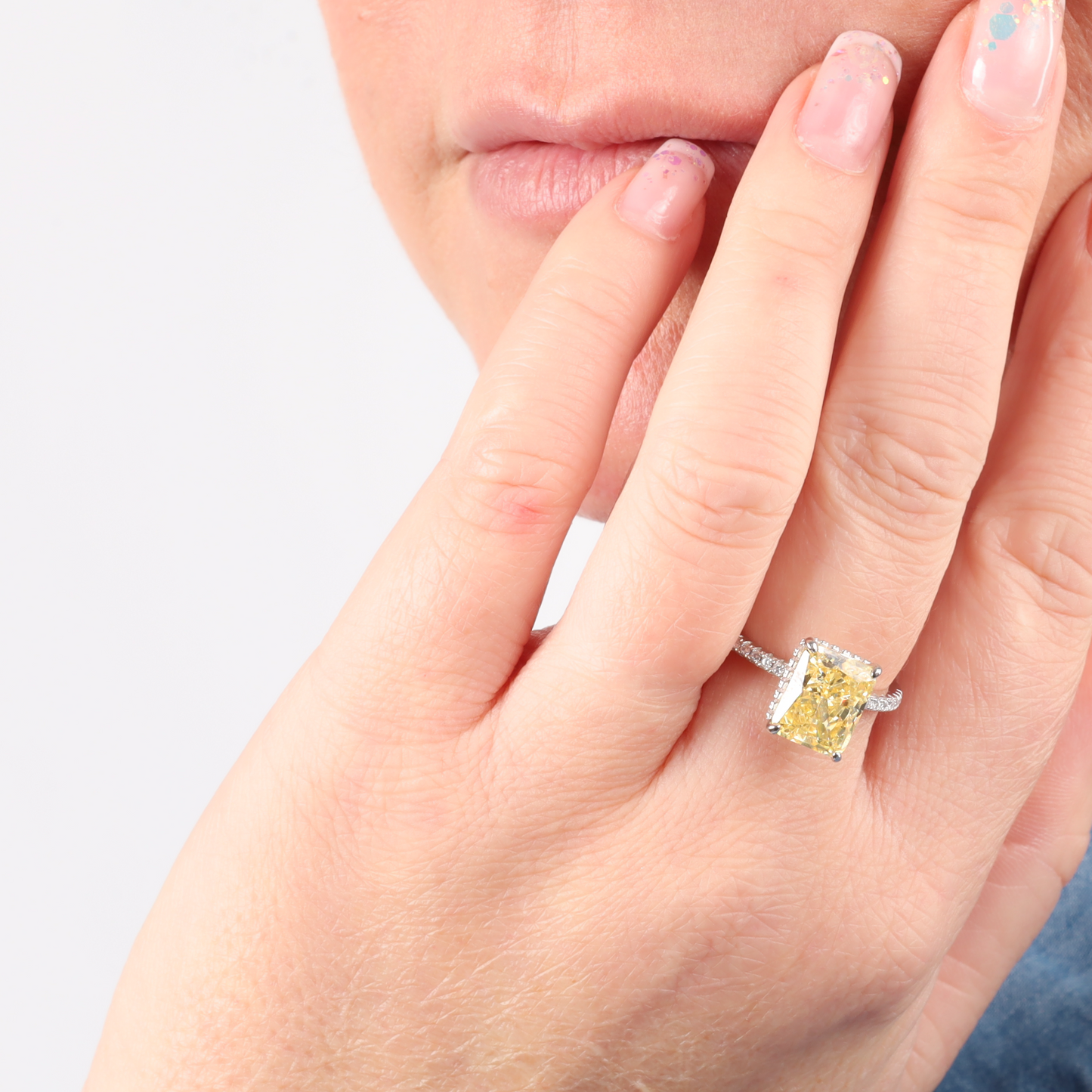 A hand graces the chin, adorned with a white gold plated Crushed Ice Yellow Radiant Promise Ring by Mia Ishaaq, dazzling in its brilliance.