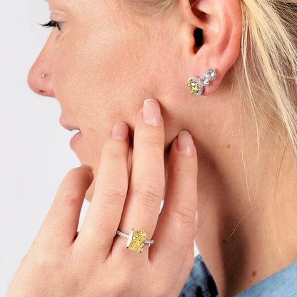 Close-up of a person showcasing diamond earrings and a stunning Crushed Ice Yellow Radiant Promise Ring by Mia Ishaaq, featuring sterling silver details, as they touch their cheek.