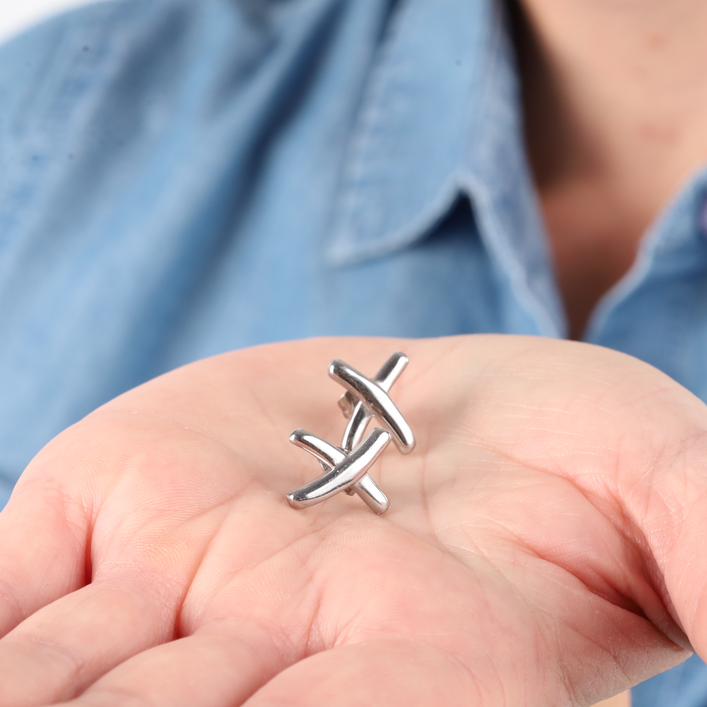 A hand holds silver metal puzzle pieces against a blue shirt backdrop, elegantly paired with the Gemma Owen Inspired XO Cross Stud Earrings in Silver by Mia Ishaaq® for a touch of subtle sophistication.