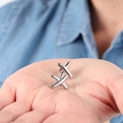 A hand holds silver metal puzzle pieces against a blue shirt backdrop, elegantly paired with the Gemma Owen Inspired XO Cross Stud Earrings in Silver by Mia Ishaaq® for a touch of subtle sophistication.