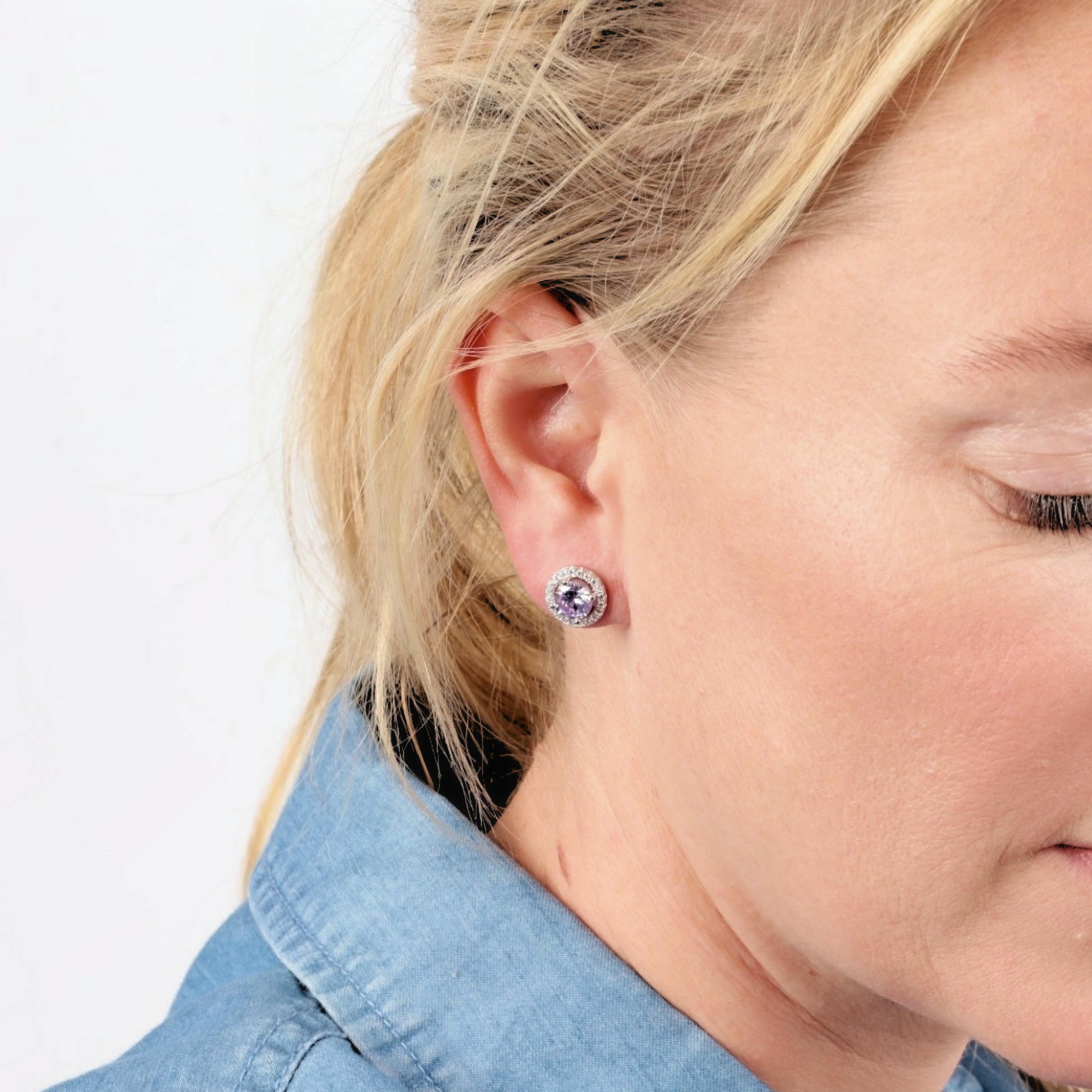 A woman with blonde hair wears a blue shirt and round, 925 sterling silver stud earrings featuring lavender CZ gems by Mia Ishaaq.