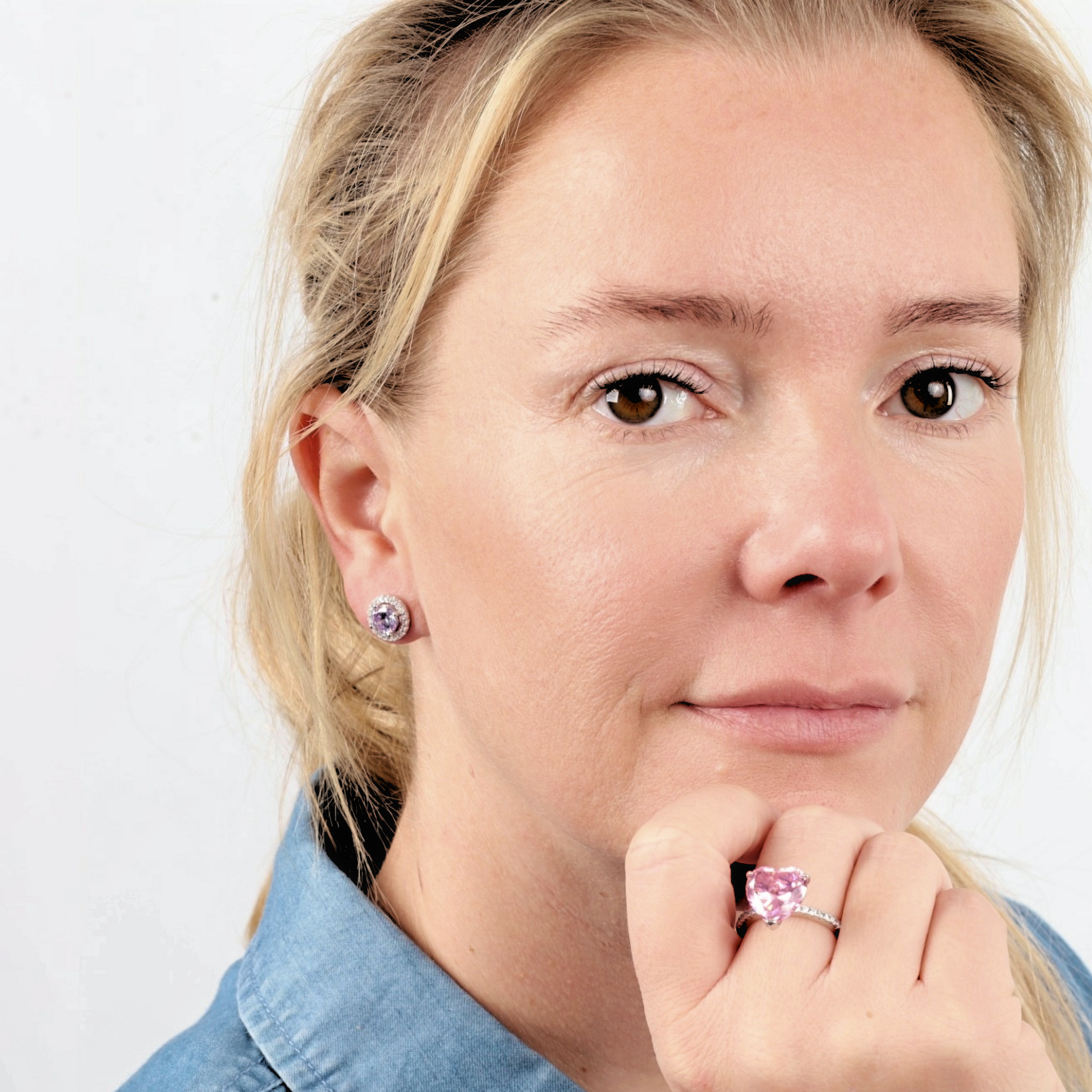 A woman with blonde hair showcases her exquisite Mia Ishaaq 925 Sterling Silver LAVENDER CZ Stud Earrings and a matching ring, both elegantly set in 925 sterling silver. She rests her chin on her hand, highlighting the sparkle of the gems against her blue shirt.