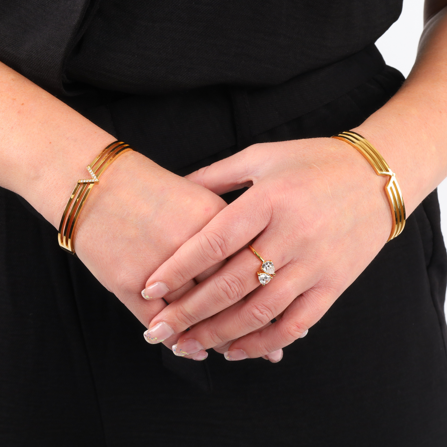 Close-up of hands adorned with a Mia Ishaaq® Gold Crystal V Line Harmony Bangle and a ring featuring a large gemstone, all elegantly plated in 18k gold. These exquisite pieces rest gracefully against a sleek black outfit, embodying victory through harmony.