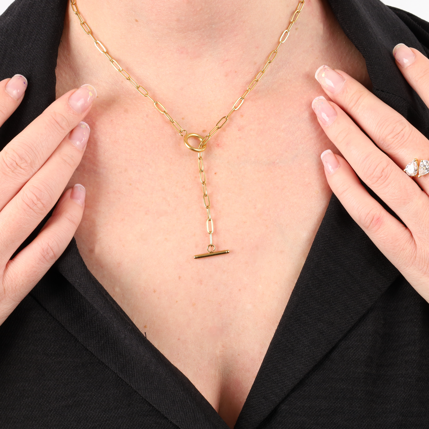 A person elegantly adorns the *Link Chain T-Bar Layering Necklace* by Mia Ishaaq, featuring a water-resistant 18k gold-plated chain with a toggle clasp, perfectly highlighting their black top as their hands rest lightly near the collar.