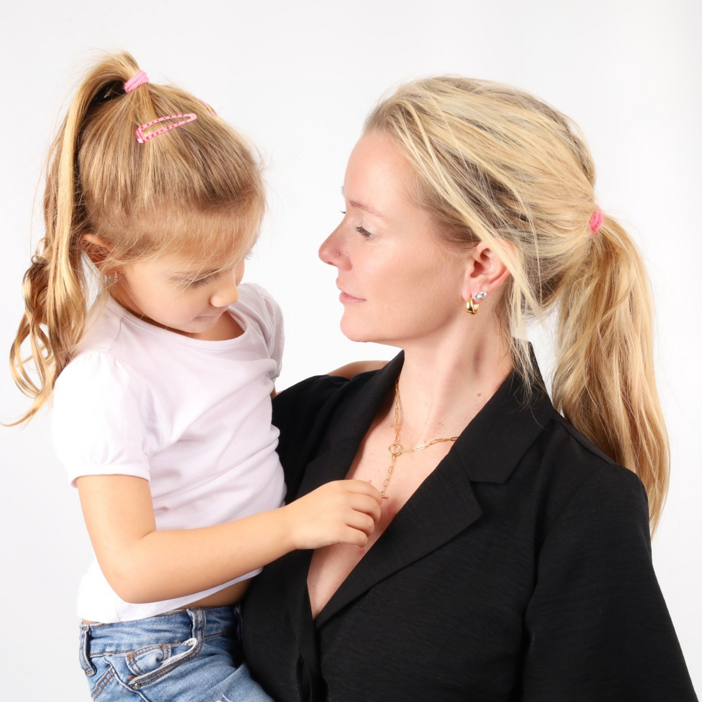 A woman in a black top, adorned with a Mia Ishaaq Link Chain T-Bar Layering Necklace, holds a young girl with a ponytail. Both are gazing at each other against a plain background, their bond as timeless as 18k gold-plated jewelry.