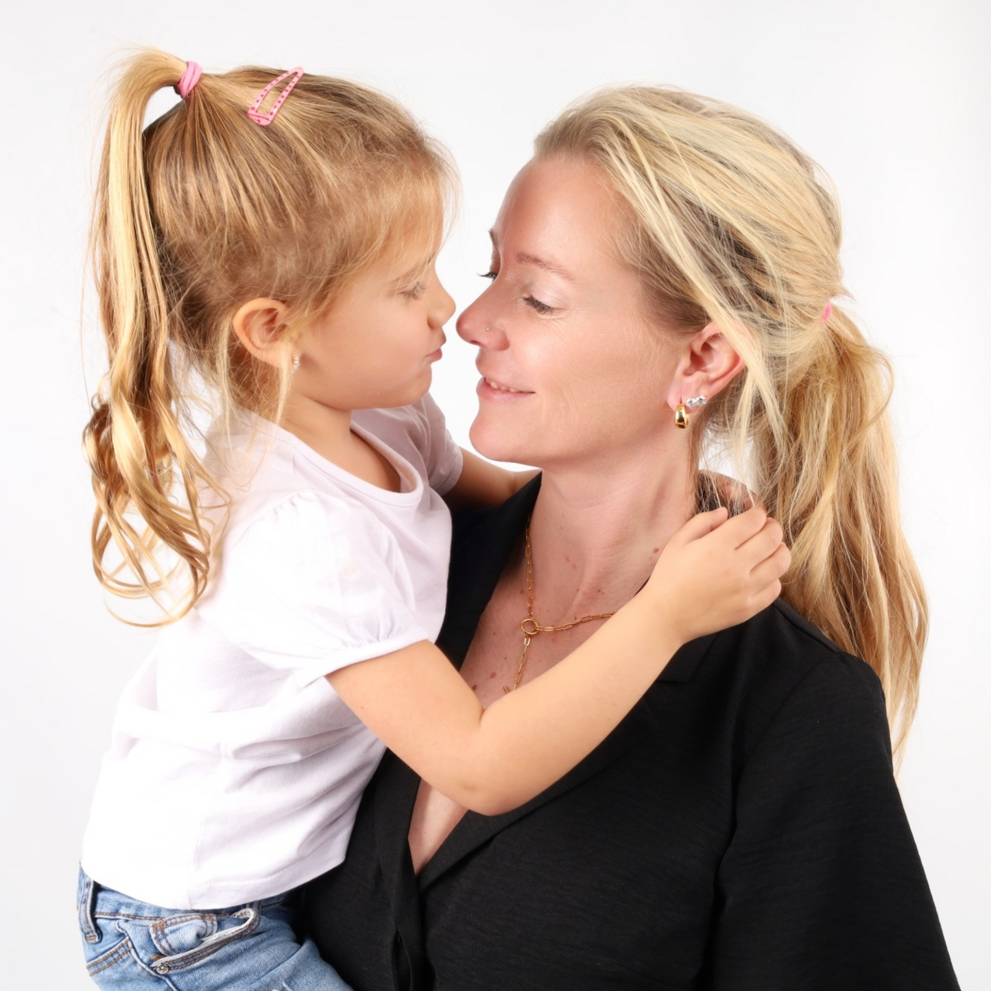 A woman in a black top holds a young girl in a white shirt, both smiling and touching noses. The Mia Ishaaq 18k gold plated, water-resistant Link Chain T-Bar Layering Necklace gleams softly, capturing the joy of the moment.