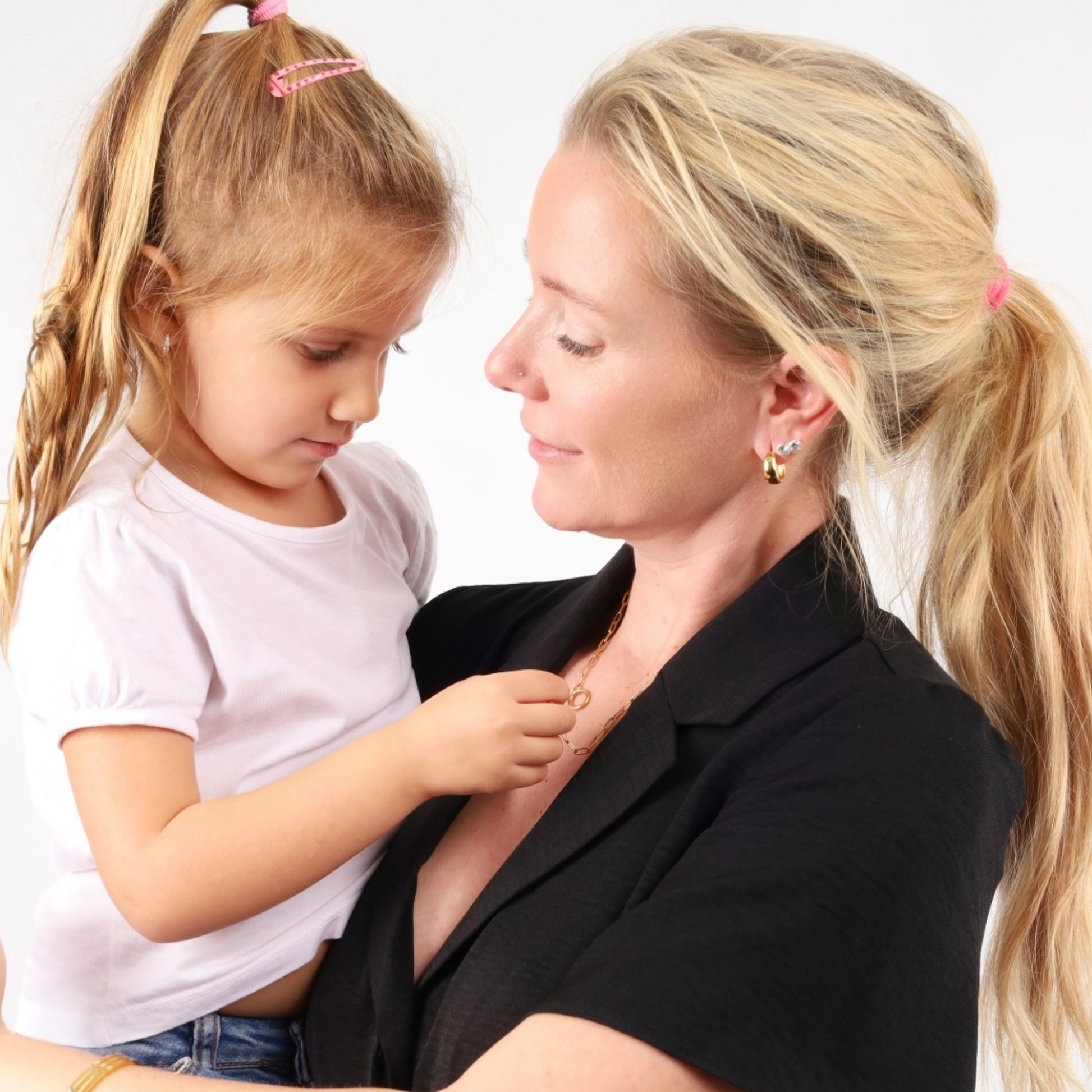 A woman lovingly holding a young girl, both with ponytails and wearing dazzling Mia Ishaaq® Gold Crystal Hoop Earrings, against a plain white background.