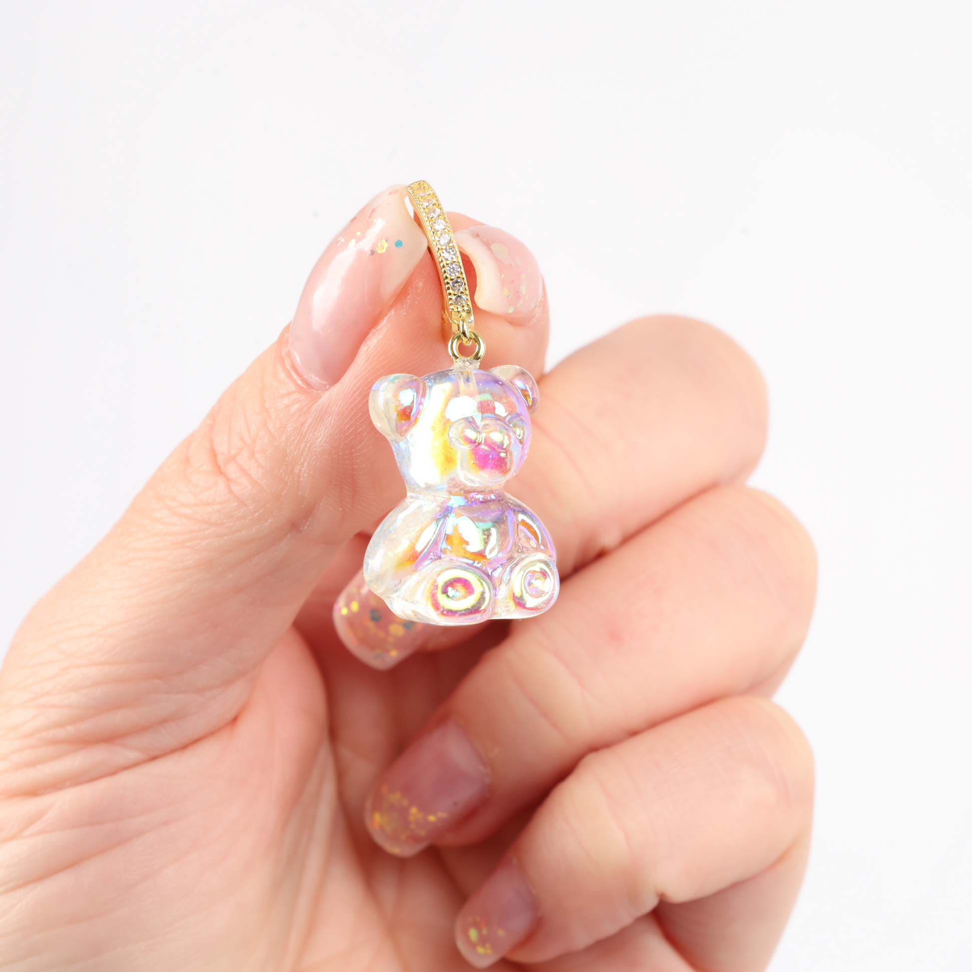 A hand holds an intricately crafted, shiny iridescent teddy bear charm featuring a delicate gold ring. This enchanting piece would pair perfectly with the Shimmering Teddy Bear Earrings by Mia Ishaaq to complete the look.