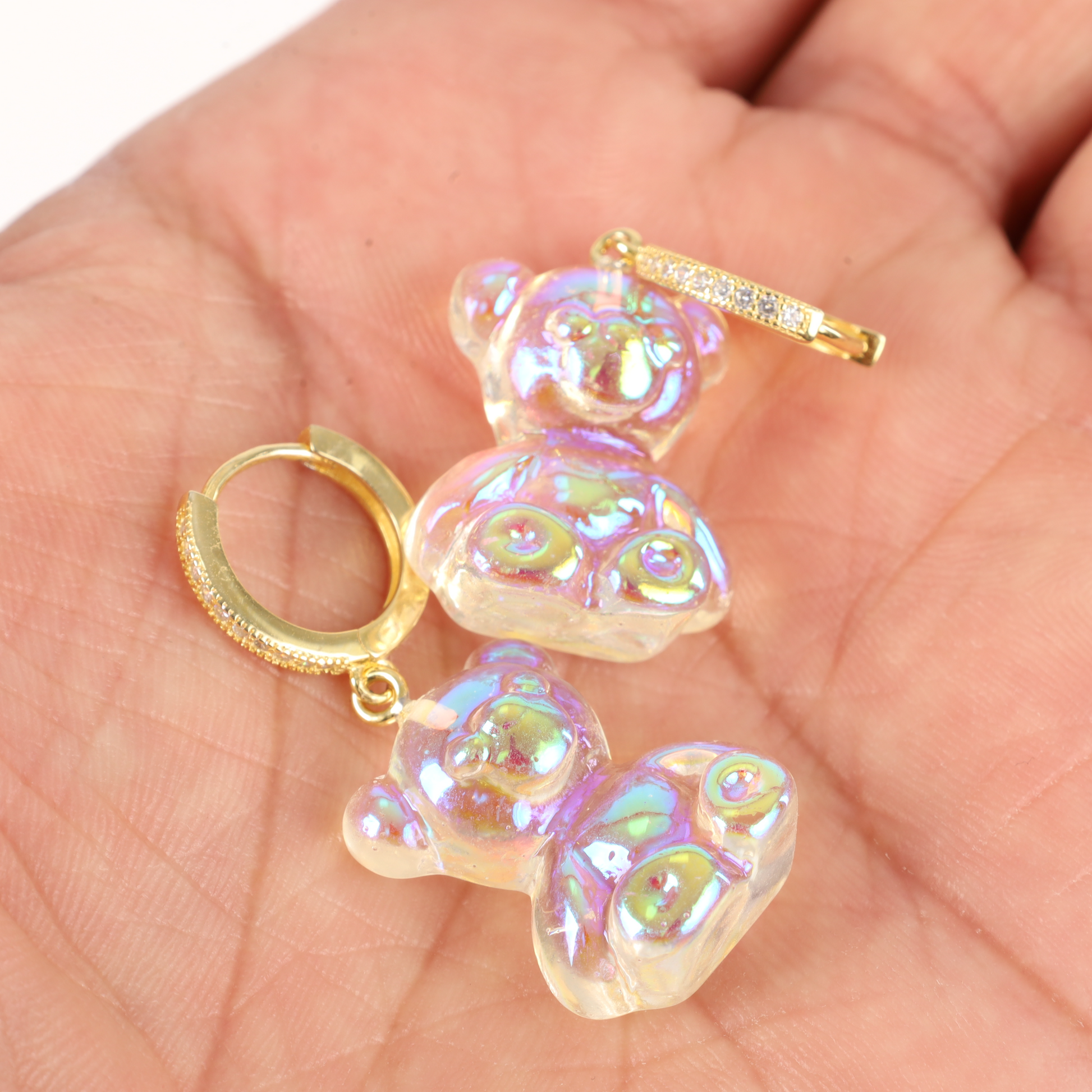 A hand holds a pair of Shimmering Teddy Bear Earrings by Mia Ishaaq, featuring iridescent teddy bears and delicate gold hoops adorned with sparkling cubic zirconia.