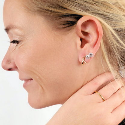 A woman with short blonde hair wears minimalistic bold accessories from Mia Ishaaq®, including the Gold Loop Cross Stud Earrings. She also sports a silver nose stud and a gold ring, smiling gently.