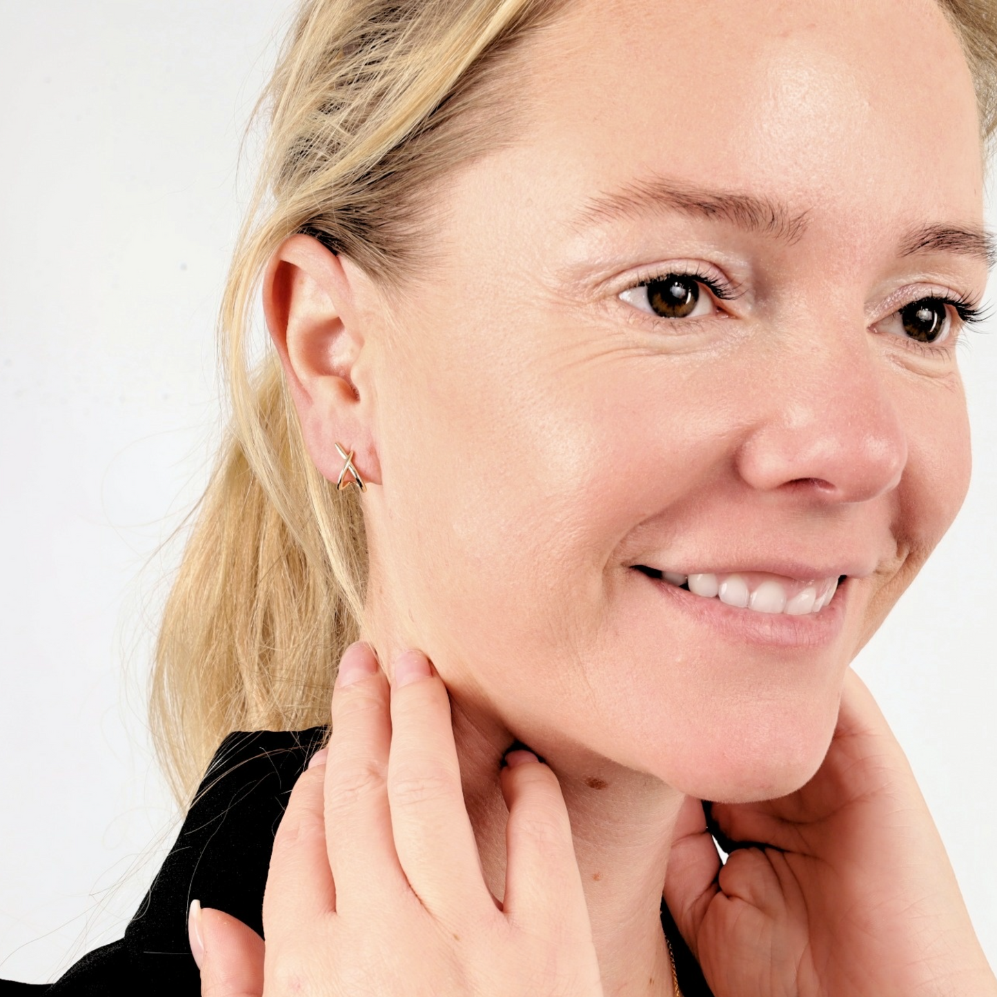 A smiling woman with blonde hair showcases Mia Ishaaq®'s Gold Loop Cross Stud Earrings in gold-plated sterling silver, gently touching her neck against a plain background.