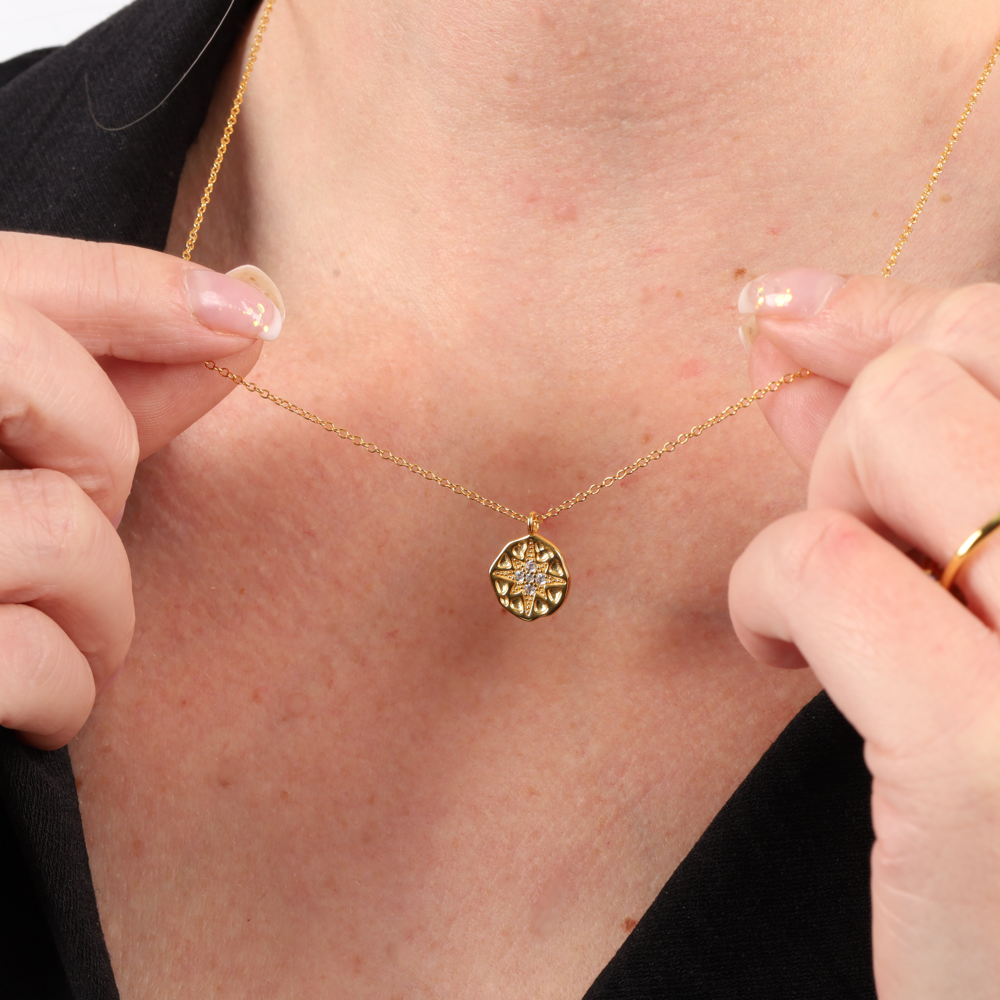 A person wearing the Polaris Coin Pendant Necklace - Gold by Mia Ishaaq®, adjusting it with both hands near a black jacket.