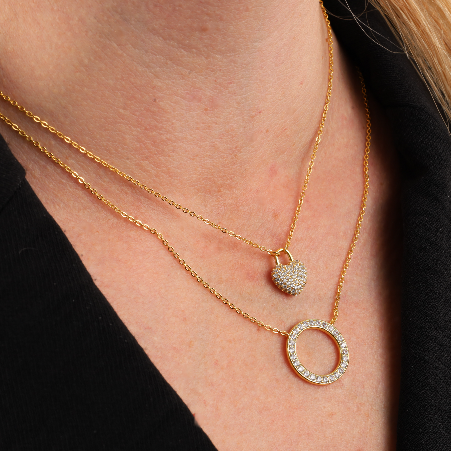 Close-up of two gold necklaces featuring a round pendant and a small heart, elegantly layered against a black shirt using the Mia Ishaaq Gold Necklace Layering Clip.