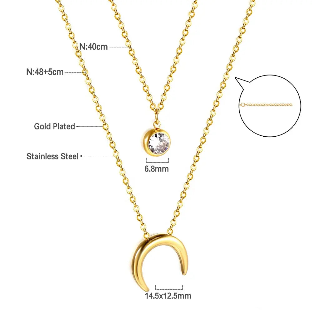 The Moon & Crystal Pendant Layered Necklace by Mia Ishaaq showcases an 18k gold-plated crescent pendant alongside a second chain adorned with a round cubic zirconia pendant. Measurements provided.